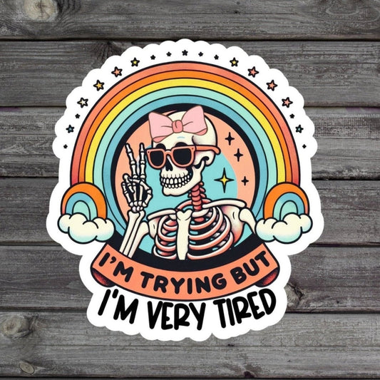 Sticker with a skull wearing a bow and the text 'I'm Tired But I'm Trying' in a cute, whimsical style